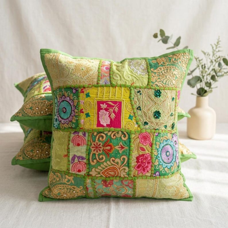 May include: A square throw pillow featuring a patchwork design of various green, yellow, and gold fabric panels. The surface is decorated with intricate embroidery, floral motifs, and small sequins in shades of pink, blue, and white.
