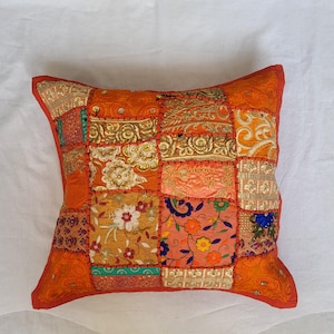 May include: A square throw pillow featuring a patchwork design of various orange, gold, and multi-colored fabric squares. The surface is decorated with intricate embroidery, floral patterns, and metallic thread accents, all joined by visible orange stitching.