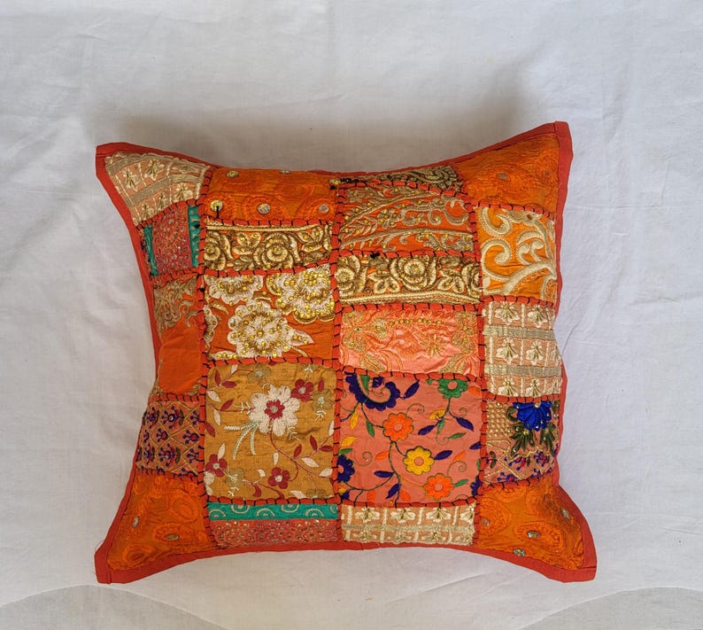 May include: A square throw pillow featuring a patchwork design of various orange, gold, and multi-colored fabric squares. The surface is decorated with intricate embroidery, floral patterns, and metallic thread accents, all joined by visible orange stitching.