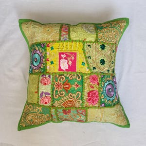 May include: A square throw pillow featuring a patchwork design of various green, yellow, and pink fabrics. The surface is decorated with intricate embroidery, floral patterns, sequins, and metallic gold thread accents, all joined by visible green stitching.