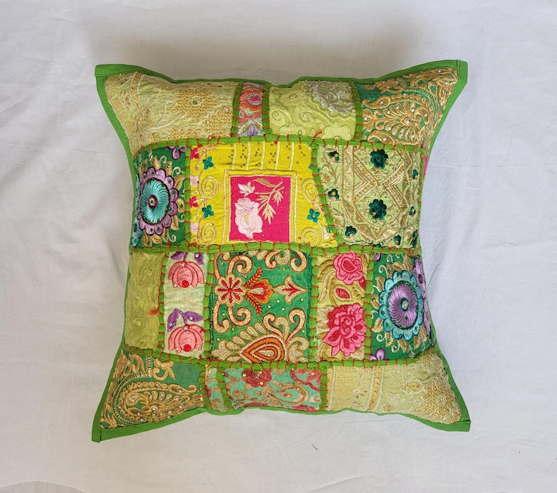 May include: A square throw pillow featuring a patchwork design of various green, yellow, and pink fabrics. The surface is decorated with intricate embroidery, floral patterns, sequins, and metallic gold thread accents, all joined by visible green stitching.