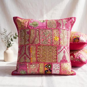 Op de afbeelding: Een vierkant sierkussen met een patchworkontwerp van diverse roze en goudkleurige stoffen. Het oppervlak is versierd met ingewikkeld metaaldraadborduurwerk, bloemmotieven, kleine ronde spiegeltjes en geometrische stiksels in magentatinten.