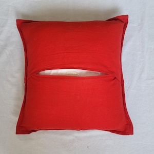 May include: A square red throw pillow cover featuring an envelope-style closure on the front. The fabric has a visible texture and a solid, vibrant red color. A small portion of a white inner pillow insert is visible through the center opening.
