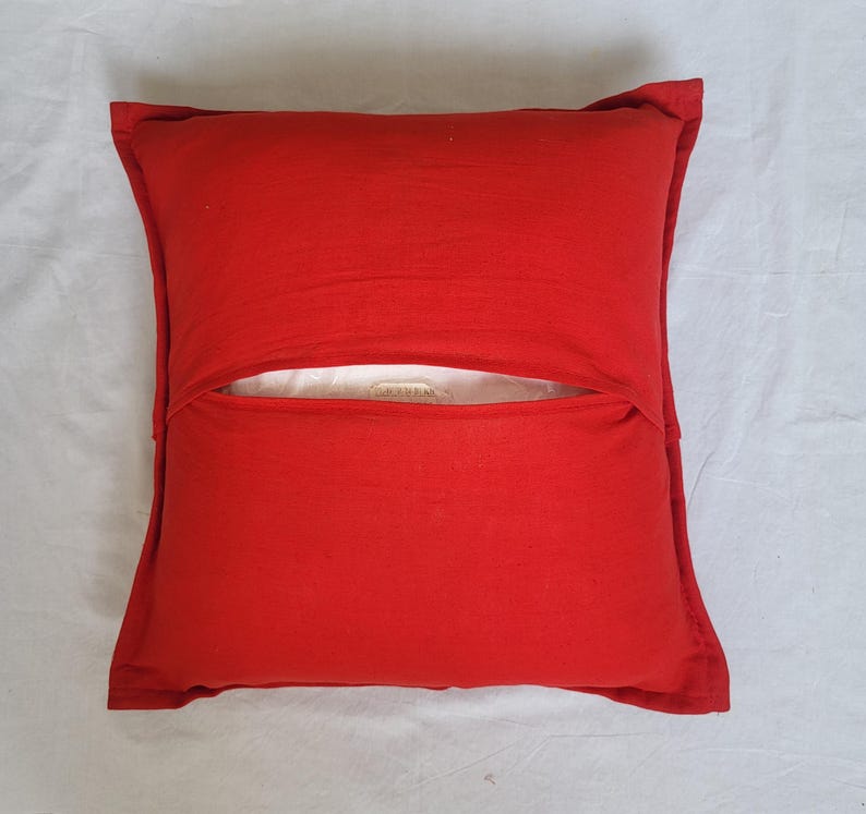 May include: A square red throw pillow cover featuring an envelope-style closure on the front. The fabric has a visible texture and a solid, vibrant red color. A small portion of a white inner pillow insert is visible through the center opening.