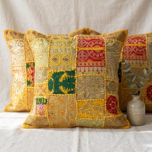 May include: A square throw pillow featuring a patchwork design of embroidered fabric panels in shades of yellow, gold, red, and green. The surface displays intricate floral and geometric patterns with visible stitching connecting each distinct textile section.