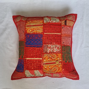 May include: A square throw pillow featuring a patchwork design of various red, orange, blue, and green fabric panels. The surface is decorated with intricate gold embroidery, metallic thread accents, small mirrors, and floral patterns throughout.