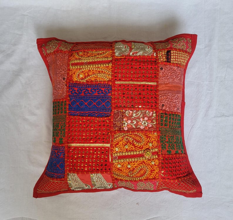 May include: A square throw pillow featuring a patchwork design of various red, orange, blue, and green fabric panels. The surface is decorated with intricate gold embroidery, metallic thread accents, small mirrors, and floral patterns throughout.