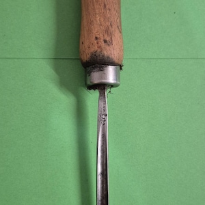 May include: A woodworking chisel with a wooden handle and a metal blade. The handle is a light brown colour, and the blade is a dark grey. The chisel is set against a green background.