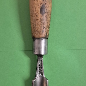 May include: A vintage wood chisel. It has a wooden handle with a metal cap and a dark grey metal blade. The handle is made of light brown wood with dark spots. The tool is set against a green background.