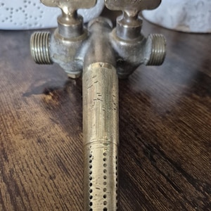 May include: Aged brass water faucet featuring two handles and a perforated lower section. The faucet exhibits signs of wear, indicating it is likely an antique or reclaimed piece. The handles are marked with the number "606".