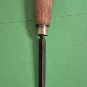 May include: A hand file with a wooden handle. The handle is light brown with dark brown rings. The file is dark grey and tapers to a point. The tool is used for shaping materials.