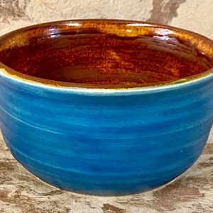 Puede incluir: Un tazón de cerámica azul con un borde marrón. El tazón tiene un acabado esmaltado y una forma ligeramente redondeada.