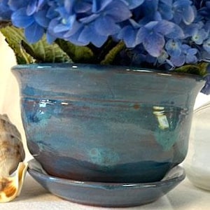 Puede incluir: Una maceta de cerámica azul verdosa con un platillo a juego. La maceta tiene una forma ligeramente redondeada y un acabado esmaltado.