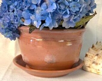 May include: A terracotta planter with a matching saucer, featuring a slightly flared rim and a smooth, glazed finish. The planter is filled with a bouquet of blue hydrangeas.