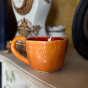 Może przedstawiać: Ręcznie wykonany ceramiczny kubek z błyszczącą pomarańczową powierzchnią zewnętrzną i ciemnoczerwonym wnętrzem. Kubek ma zaokrąglony, nieregularny kształt z grubym, zakrzywionym uchwytem po lewej stronie. Powierzchnia ma teksturę i połysk.