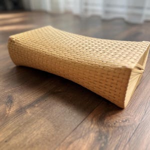Può includere: Un cuscino rettangolare intrecciato di colore beige chiaro. Il cuscino ha una forma curva ed è realizzato in un materiale a trama fitta. È posto su un pavimento di legno.