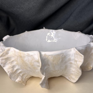 Puede incluir: Un cuenco de cerámica con interior blanco y un exterior ondulado similar a una hoja de col, con un esmalte en tonos beige pálido y crema. El cuenco tiene un borde circular ancho y abierto, con una forma orgánica y texturizada.