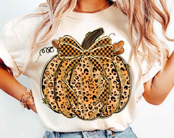 May include: Cream-colored t-shirt featuring a large pumpkin design. The pumpkin is orange with a leopard print pattern and skull accents. A checkered bow and stem complete the festive Halloween design.
