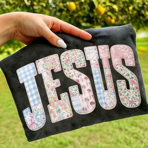 May include: A dark grey fabric item displaying the word "JESUS" in large, appliquéd letters. The letters are crafted from diverse floral and gingham fabrics, including a light blue gingham "J" and a pink floral "S".