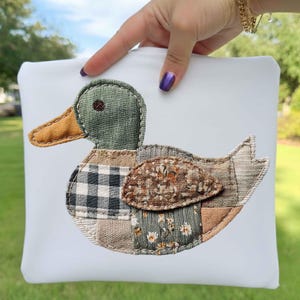 May include: A white square cushion featuring an appliqued duck design. The duck has a green head, orange beak, and a patchwork body made of various patterned fabrics in green, brown, and cream. The cushion is held up by a hand.
