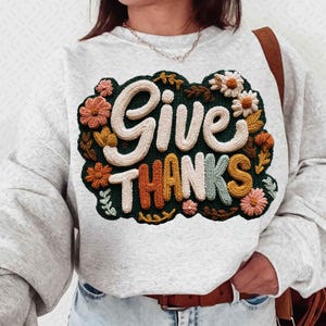 May include: A light gray sweatshirt with the words "GIVE THANKS" in a 3D embroidered design. The design features a dark green background with cream-colored lettering and colorful floral accents in shades of orange, pink, and yellow.