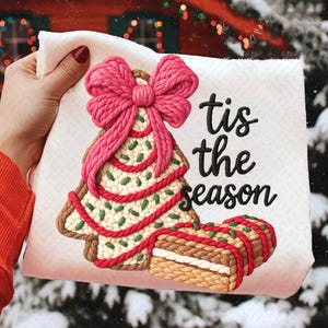 Peut inclure: Décoration de Noël en tissu blanc avec un motif de sapin en pain d'épices texturé, un nœud rose et des détails rouges et verts. Le texte "tis the season" est écrit en script noir.