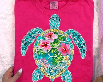 May include: A bright pink short-sleeve t-shirt featuring a large sea turtle graphic on the front. The turtle is decorated with turquoise and gold rhinestones, with a center shell design of pink hibiscus flowers and green palm leaves.