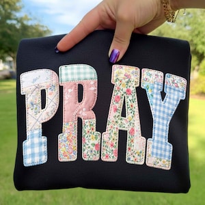 May include: Black fabric item with the word "PRAY" appliqued in colorful patchwork letters. The letters feature floral, gingham, and patterned fabrics. The item is held up against a green background.
