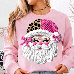 Könnte beinhalten: Rosa Sweatshirt mit einem Santa Claus-Gesicht-Design. Das Santa-Gesicht hat eine rosa Nase und Wangen, einen weißen Bart und einen rosa und Leopardenmuster-Hut. Rosa Schleifen schmücken Hut und Bart. Ein festliches Kleidungsstück zum Thema Feiertage.