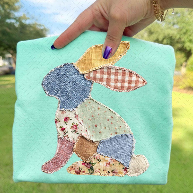May include: A light teal square pillow with a patchwork bunny design. The bunny is made of various patterned fabrics, including floral, gingham, and solid colors. The pillow is held by a hand with purple nail polish.