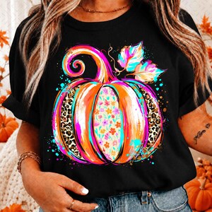 Könnte beinhalten: Ein schwarzes T-Shirt mit einer großen, farbenfrohen Kürbisgrafik. Das Kürbisdesign enthält leuchtend orangefarbene, pinke und türkisfarbene Pinselstriche, Abschnitte mit Leopardenmuster und ein mittleres Feld mit kleinen Blumenmotiven.