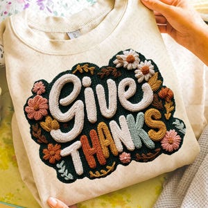 May include: Cream-colored sweatshirt with an embroidered design that reads "GIVE THANKS." The design features white, orange, and blue lettering, surrounded by floral and leaf accents in shades of pink, brown, and green. The sweatshirt is held by a person.