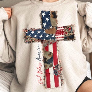 May include: A cream-colored sweatshirt featuring a cross design made from patches of fabric with patriotic patterns. The cross incorporates elements of the American flag, including stars and stripes. The words "God Bless America" are written in red.