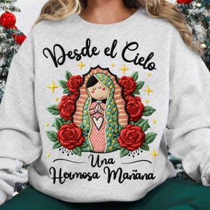 Peut inclure: Un sweat-shirt gris clair avec un motif brodé. Le motif comprend une figure centrale avec un cœur, entourée de roses rouges, de croix dorées et du texte "Desde el Cielo" et "Una Hermosa Mañana" en écriture noire.