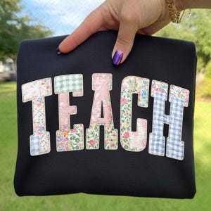 Może przedstawiać: Czarna torba typu tote z napisem "TEACH" naszytym z patchworku. Litery są w stylu uniwersyteckim, a każda z nich ma inny wzór kwiatowy lub w kratkę w odcieniach różu, błękitu i zieleni.