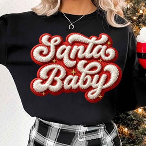 May include: Black sweatshirt with the embroidered text "Santa Baby" in red and white, accented with gold stars. The design has a retro, festive aesthetic, perfect for the holiday season.