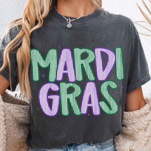 Puede incluir: Camiseta gris oscuro con las palabras "MARDI GRAS" en letras grandes, verdes y moradas con rayas verticales. La camiseta tiene cuello redondo y mangas cortas. El texto est&aacute; delineado en morado y verde, con estrellas.