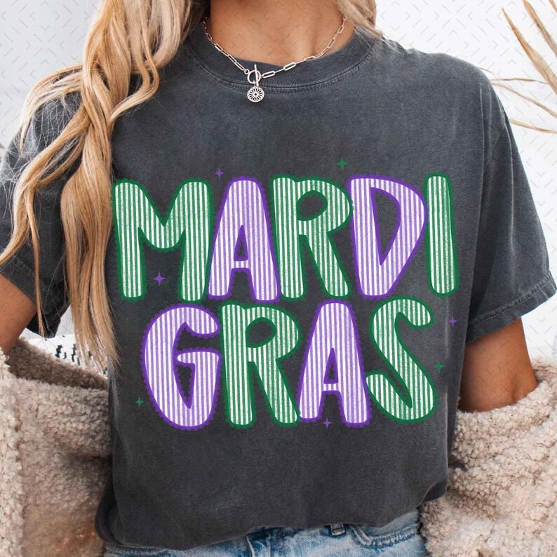 Puede incluir: Camiseta gris oscuro con las palabras "MARDI GRAS" en letras grandes, verdes y moradas con rayas verticales. La camiseta tiene cuello redondo y mangas cortas. El texto est&aacute; delineado en morado y verde, con estrellas.