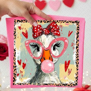May include: A pink square canvas print featuring a whimsical goose wearing pink glasses and a red bow. The goose is blowing a pink bubble and surrounded by red hearts. The border has a leopard print design.
