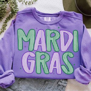 Puede incluir: Una sudadera lila con la inscripci&oacute;n "MARDI GRAS" en letras a rayas verdes y blancas. Las letras est&aacute;n delineadas en verde, con detalles de estrellas. La sudadera est&aacute; doblada sobre una superficie blanca, con un sombrero verde visible.
