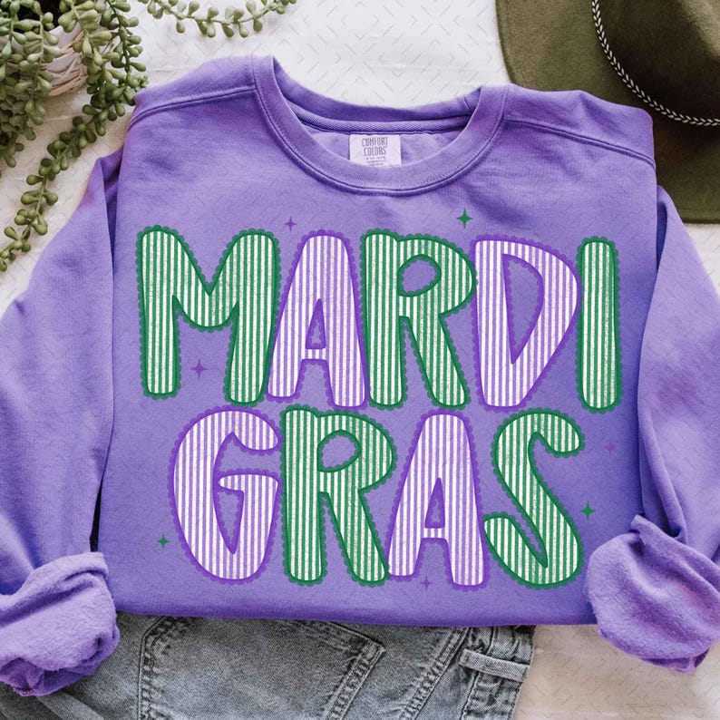 Puede incluir: Una sudadera lila con la inscripci&oacute;n "MARDI GRAS" en letras a rayas verdes y blancas. Las letras est&aacute;n delineadas en verde, con detalles de estrellas. La sudadera est&aacute; doblada sobre una superficie blanca, con un sombrero verde visible.