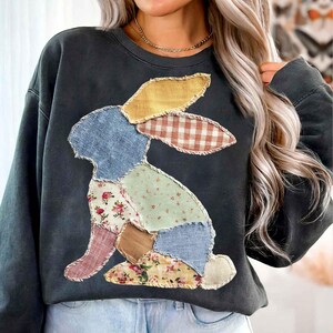 May include: Dark gray sweatshirt featuring a patchwork bunny design. The bunny is composed of various fabric patterns, including floral, gingham, and solid colors. The sweatshirt has a crew neck and long sleeves, perfect for casual wear.