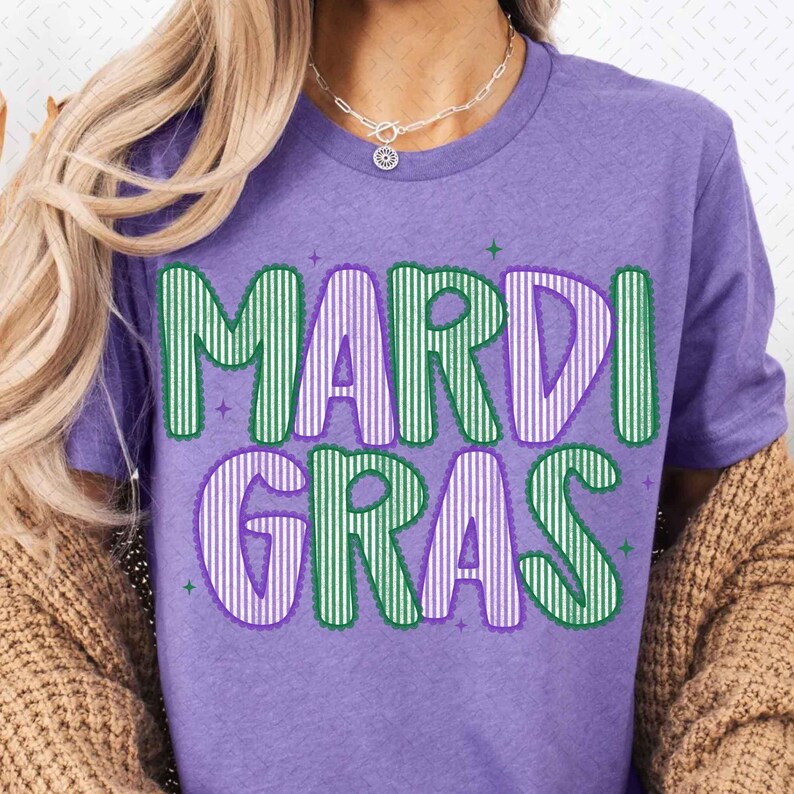 Puede incluir: Una camiseta morada con las palabras "MARDI GRAS" en letras grandes, verdes y moradas con rayas verticales. Las letras tienen un contorno blanco y est&aacute;n decoradas con peque&ntilde;as estrellas verdes. La camiseta es de color morado liso.