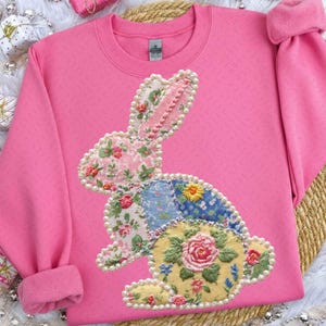 Può includere: Una felpa rosa con un design a coniglio patchwork. Il coniglio è decorato con motivi floreali, perle e ricami. La felpa ha polsini a coste e girocollo. L'estetica generale è femminile e a tema primaverile.