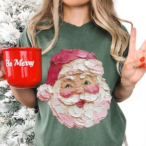 Könnte beinhalten: Ein grünes T-Shirt mit einem strukturierten Weihnachtsmann-Gesicht, komplett mit roter Mütze und weißem Bart. Eine rote Tasse mit den Worten "Be Merry" wird in der linken Hand gehalten. Das Bild hat ein festliches Urlaubs-Thema.