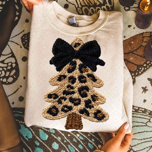 May include: Cream-colored sweatshirt featuring a knitted Christmas tree design. The tree has a leopard print pattern with a black bow at the top. The tree trunk is brown. The sweatshirt is folded and held by hands.