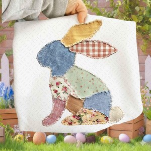 May include: A white square fabric with a patchwork bunny design. The bunny is made of various patterned fabrics in blue, yellow, red, and floral prints. The background has a subtle white pattern. The image is held up by a person.