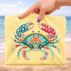 May include: A yellow tote bag with a detailed crab design. The crab is decorated with colourful gemstones in shades of blue, pink, and turquoise. The crab's body is teal with a floral design. The background is a beach scene.