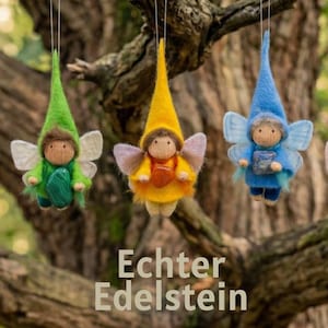 May include: Five whimsical felt fairy ornaments with pointed hats and delicate wings hang from a tree branch. Each fairy is a different color: pink, green, yellow, blue, and burgundy. The text "Echter Edelstein" is visible.