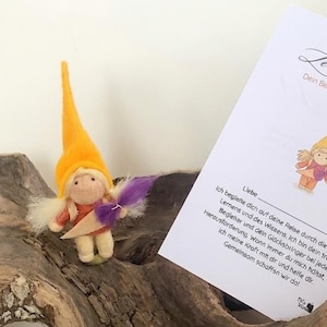 Peut inclure: Une figurine de gnome en feutre faite à la main, avec un chapeau jaune et des ailes violettes, accompagnée d'une carte en allemand. La carte présente un texte et une illustration de gnome. Le tout est posé sur du bois flotté.